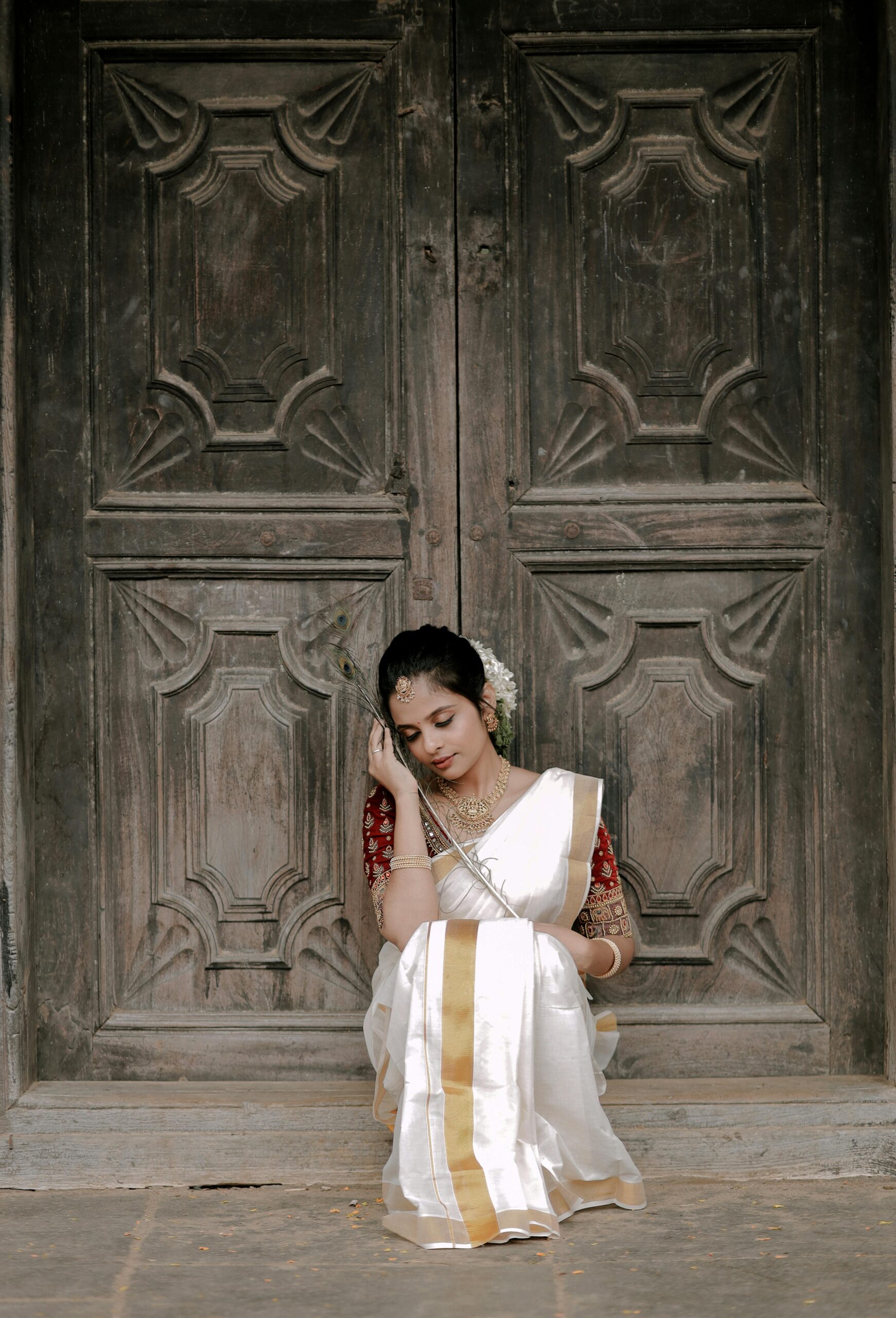 A woman in a white and gold saree sitting pensively by an intricately carved door.